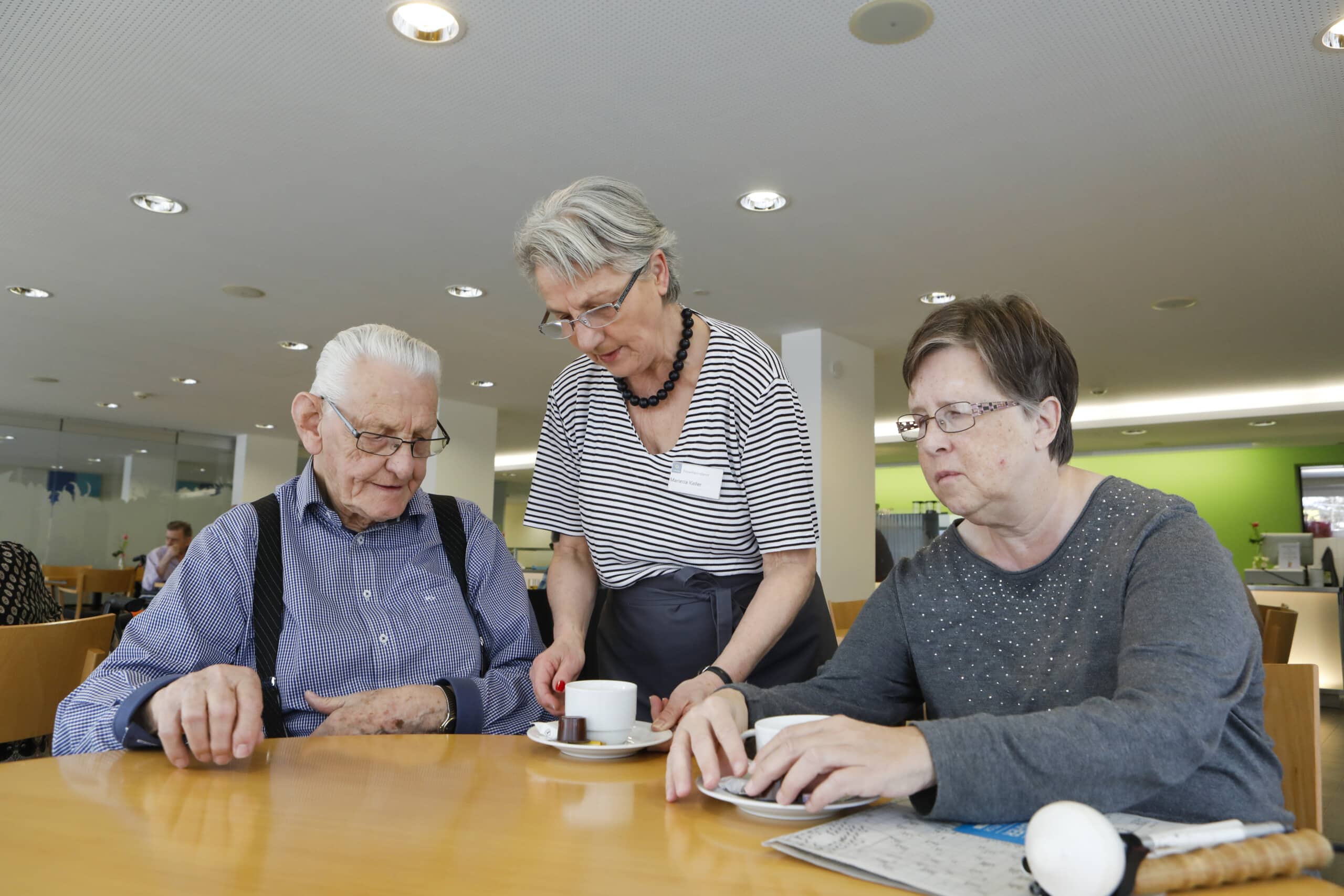 Ein älterer Mann und eine ältere Frau sitzen an einem Tisch, während ein Mitarbeiter in einem gut beleuchteten Gemeinschaftsspeisesaal Kaffee serviert.