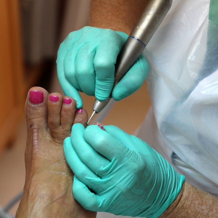 Eine Person mit grünen Handschuhen behandelt die Fußnägel einer anderen Person mit einem Handgerät mit rosa Nagellack.