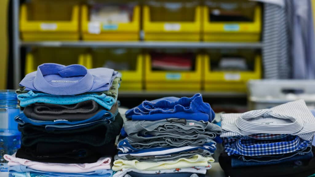 Stapel gefalteter Kleidung in verschiedenen Farben und Mustern sind auf einem Tisch angeordnet, im Hintergrund sind gelbe Aufbewahrungsbehälter zu sehen.