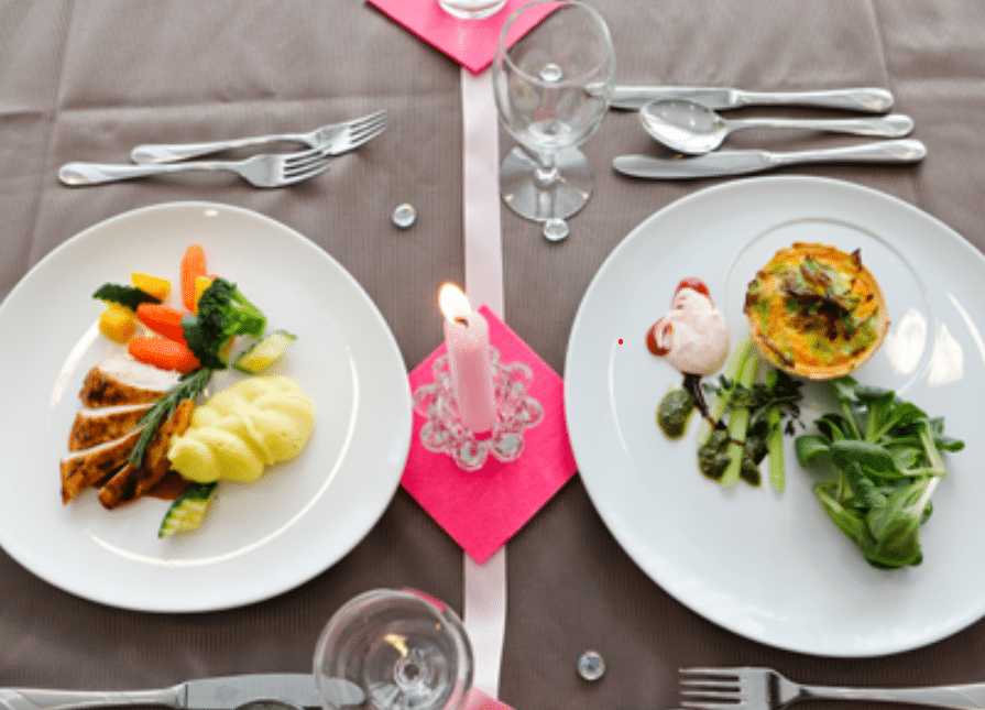 Zwei Teller mit Gourmet-Essen auf einem förmlich gedeckten Tisch mit einer brennenden rosa Kerze in der Mitte, Gläser und Besteck ordentlich auf einer grauen Tischdecke angeordnet.