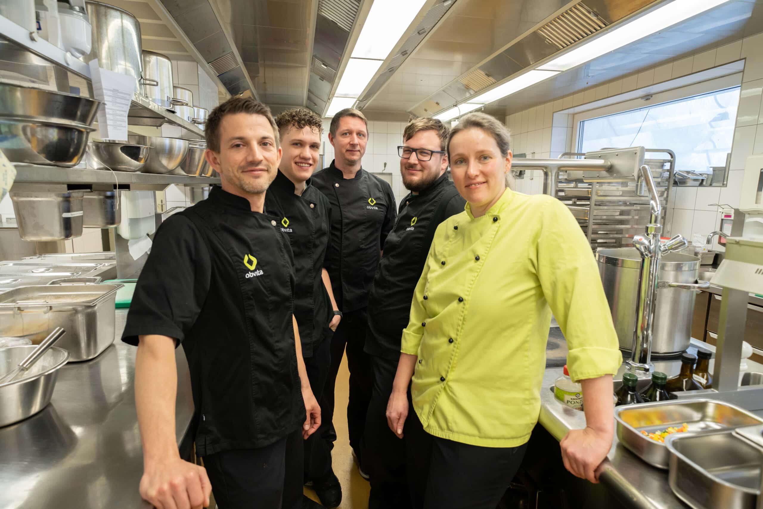 Fünf Köche in Uniform stehen zusammen in einer Großküche, umgeben von Geräten und Theken aus Edelstahl.