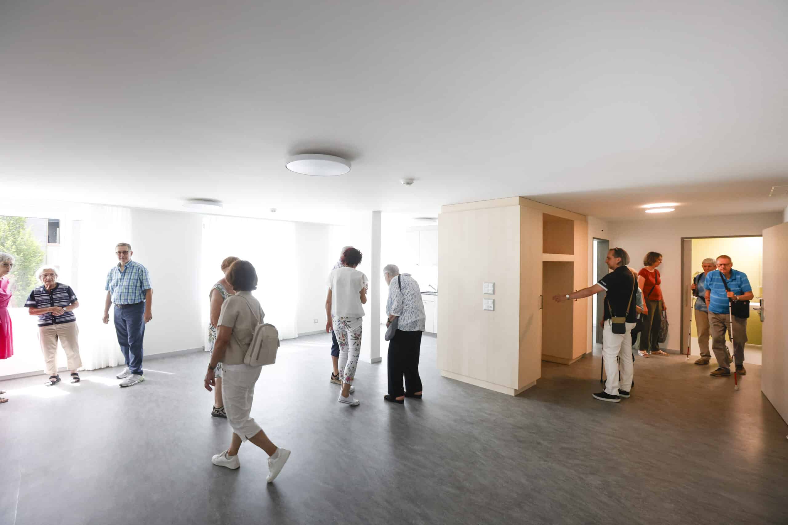Eine Gruppe von Menschen besichtigt einen hellen, unmöblierten Raum mit großen Fenstern und hellen Wänden und Böden.