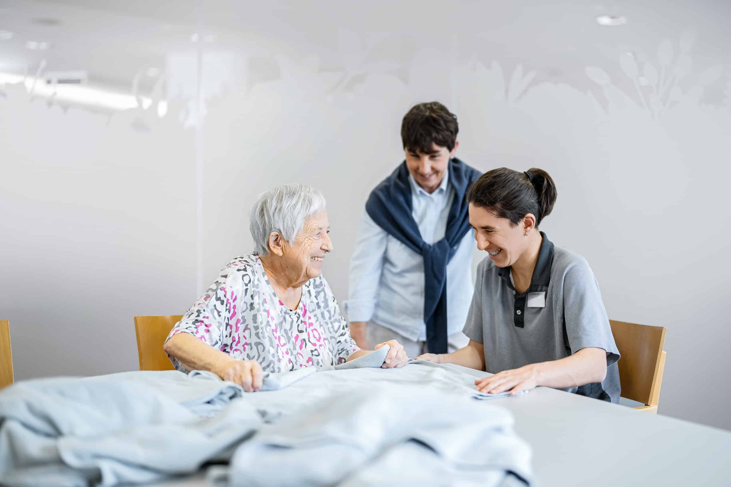 Eine ältere Frau und zwei Pflegekräfte sitzen an einem Tisch, falten gemeinsam Wäsche, lächeln und unterhalten sich in einem hellen, sauberen Raum.