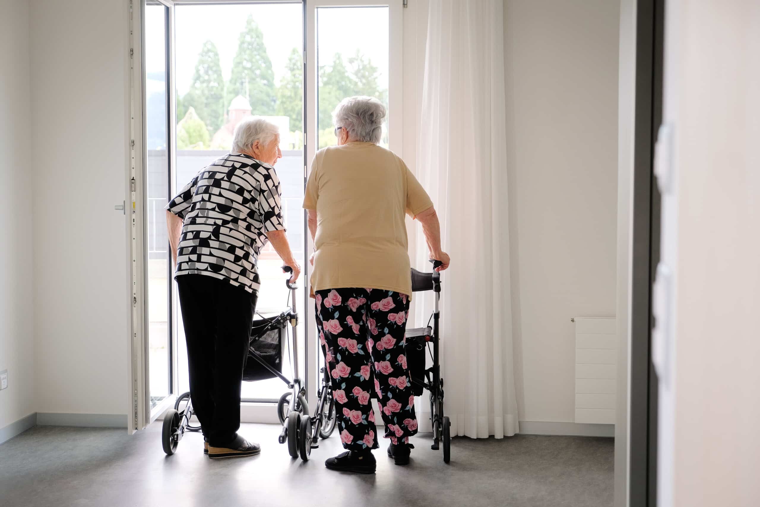 Zwei ältere Frauen mit Gehhilfen stehen nebeneinander vor einer offenen Tür und blicken aus einem hellen, minimalistischen Raum nach draußen.