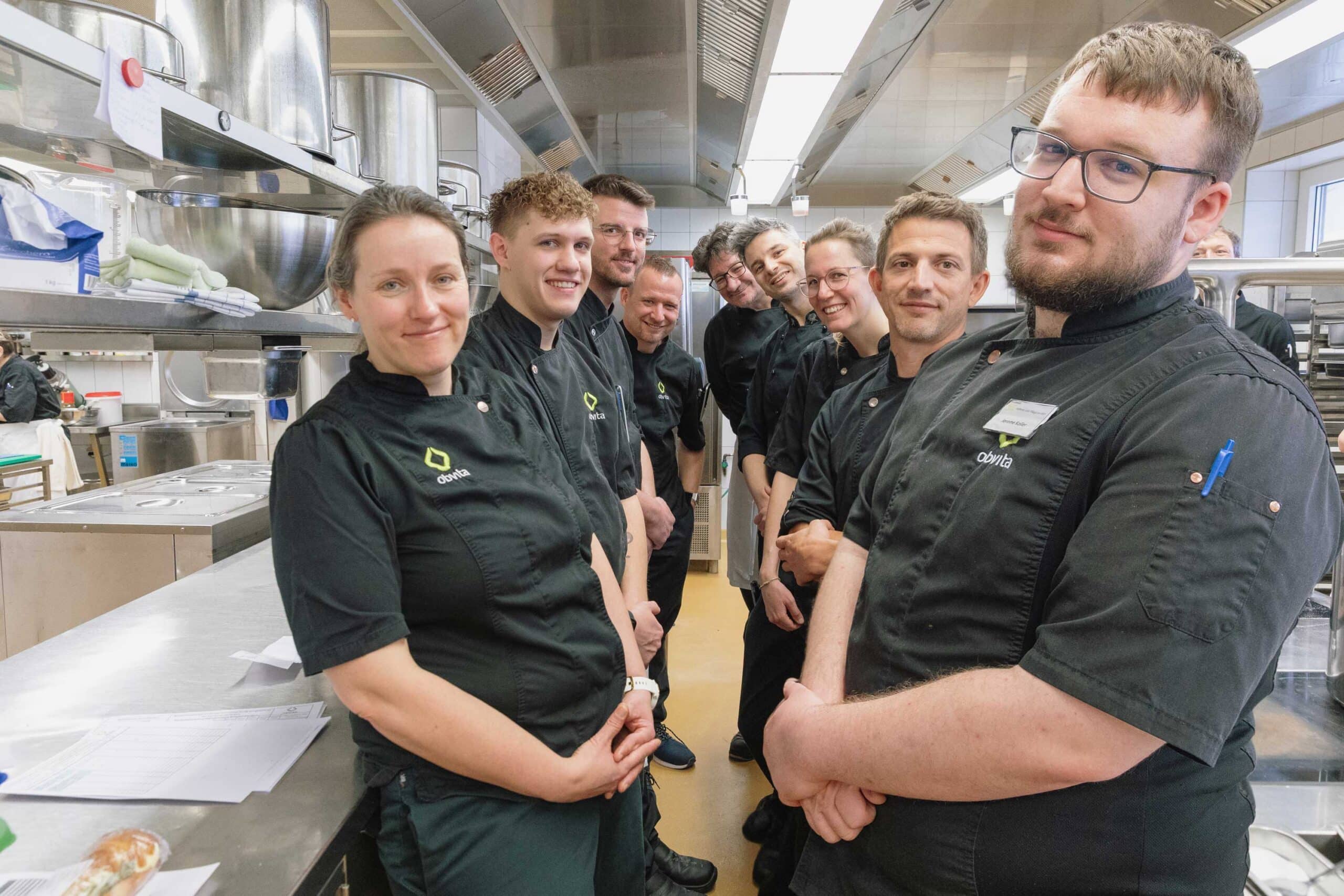 Eine Gruppe von Köchen in schwarzen Uniformen steht zusammen in einer Großküche und posiert für ein Foto neben Edelstahltheken und -geräten.