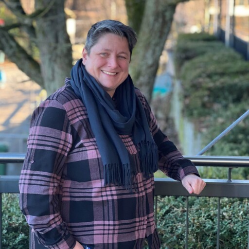 Person mit kurzen Haaren, die ein rosa kariertes Hemd und einen dunklen Schal trägt, steht im Freien an einem Geländer mit Bäumen und Grünpflanzen im Hintergrund.
