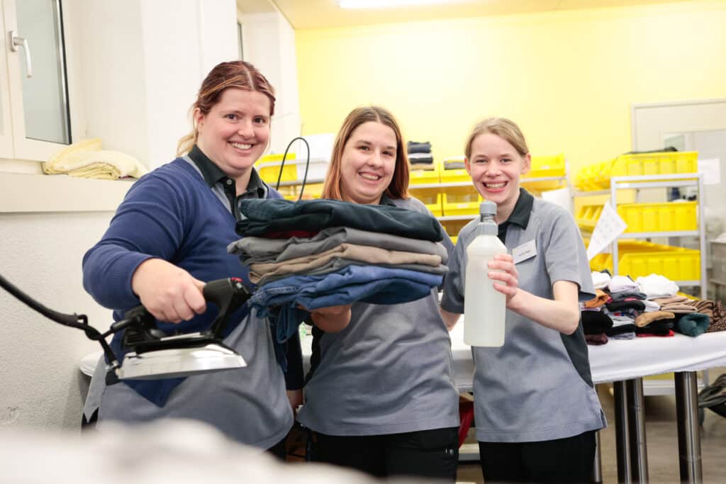 Drei Frauen in Uniform stehen in einer Waschküche, eine mit einem Bügeleisen, eine mit gefalteter Wäsche und die dritte mit einer Flasche, im Hintergrund Regale mit gelben Behältern.
