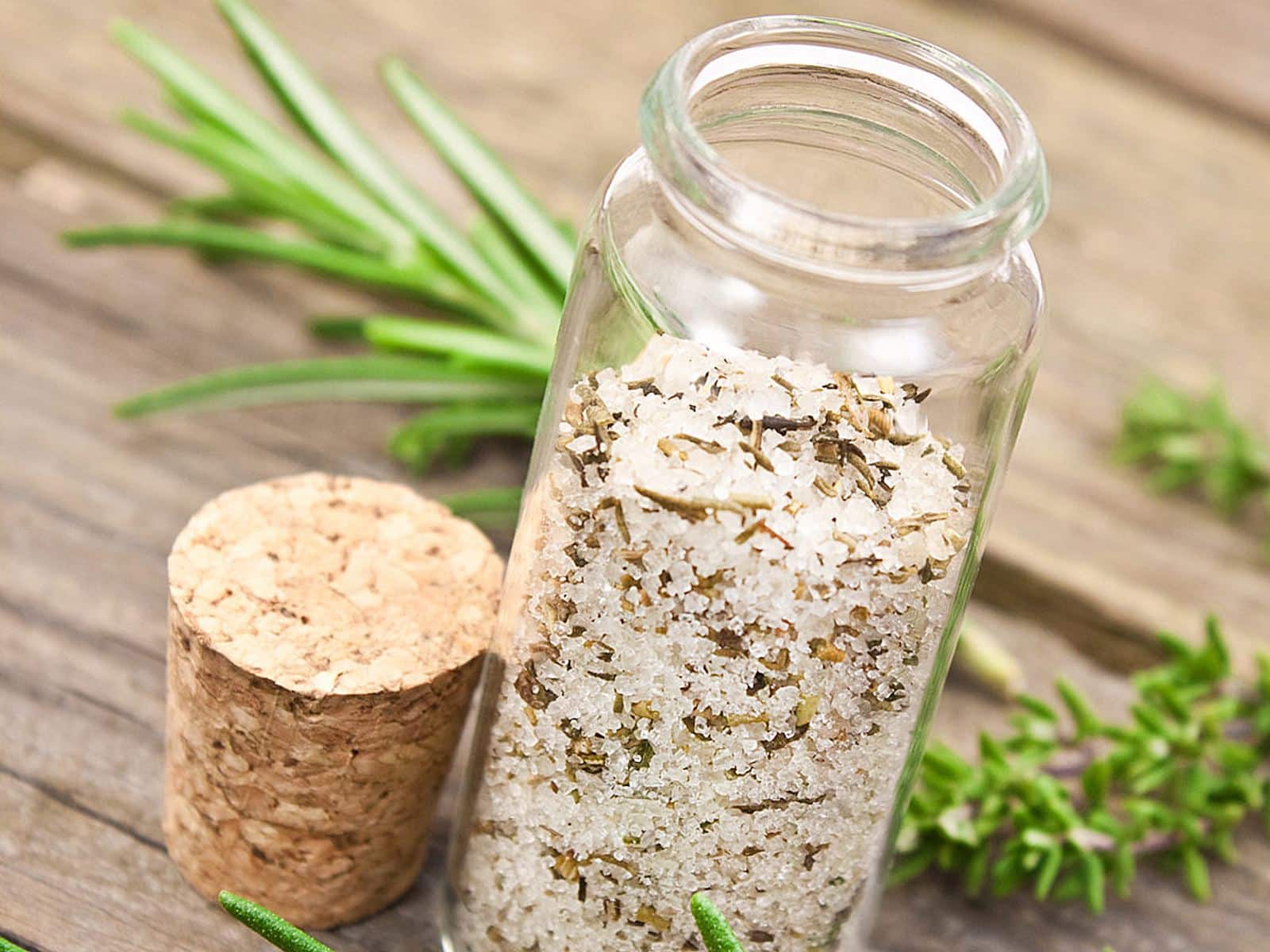 Mit grobem Salz und Kräutern gefülltes Glasgefäß, das auf der Seite liegt, mit einem Korken daneben und frischen Rosmarinzweigen im Hintergrund auf einer Holzfläche.