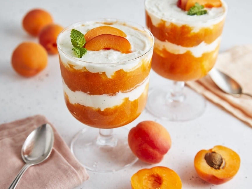 Zwei Gläser mit einem Dessert aus geschichteten Aprikosen und Sahne, garniert mit Aprikosenscheiben und Minze. Ganze und halbierte Aprikosen, Löffel und eine Serviette stehen auf dem Tisch.