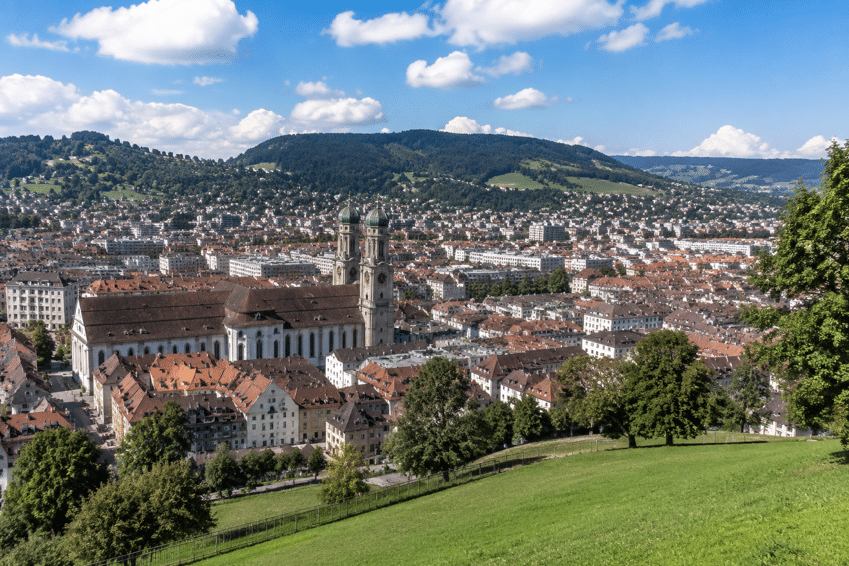Panoramablick auf eine europäische Stadt mit einer großen Kirche, Gebäuden mit roten Dächern und Hügeln im Hintergrund unter einem teilweise bewölkten Himmel.