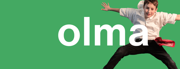 Ein Junge in einem weißen Hemd und einer schwarzen Hose springt spielerisch vor einem grünen Hintergrund, auf dem in großen weißen Buchstaben das Wort „olma“ steht.